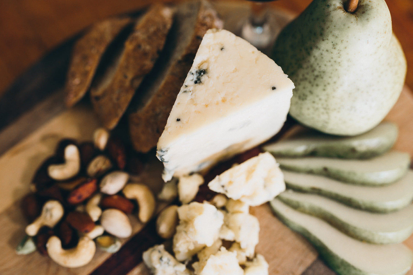 Bleu Claire cheese is shown on a wooden board with salted nuts, seedy bread, and thinly sliced pear. The cheese has a gentle blue veining.