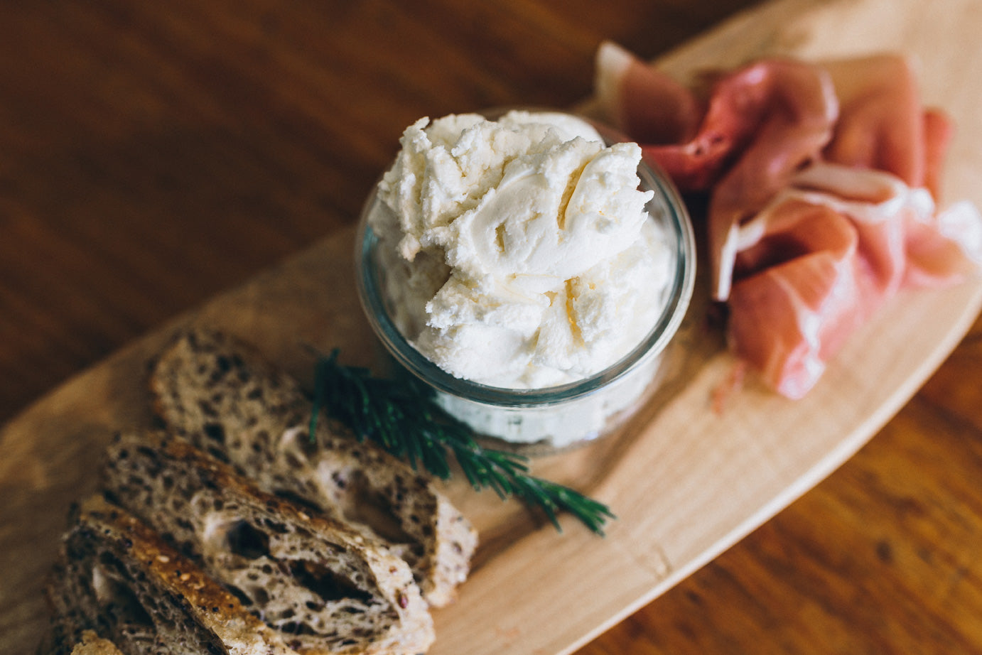 Fromage Frais natural is styled on a wooden board next to prosciutto and a rustic bread. The cheese is spooned into a serving jar for display.