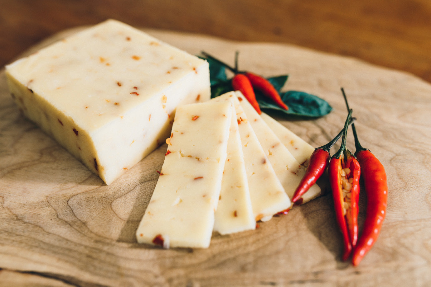 Hot Jill cheese is shown on a wooden board next to fresh chili peppers. The cheese is flecked with red chili flakes.