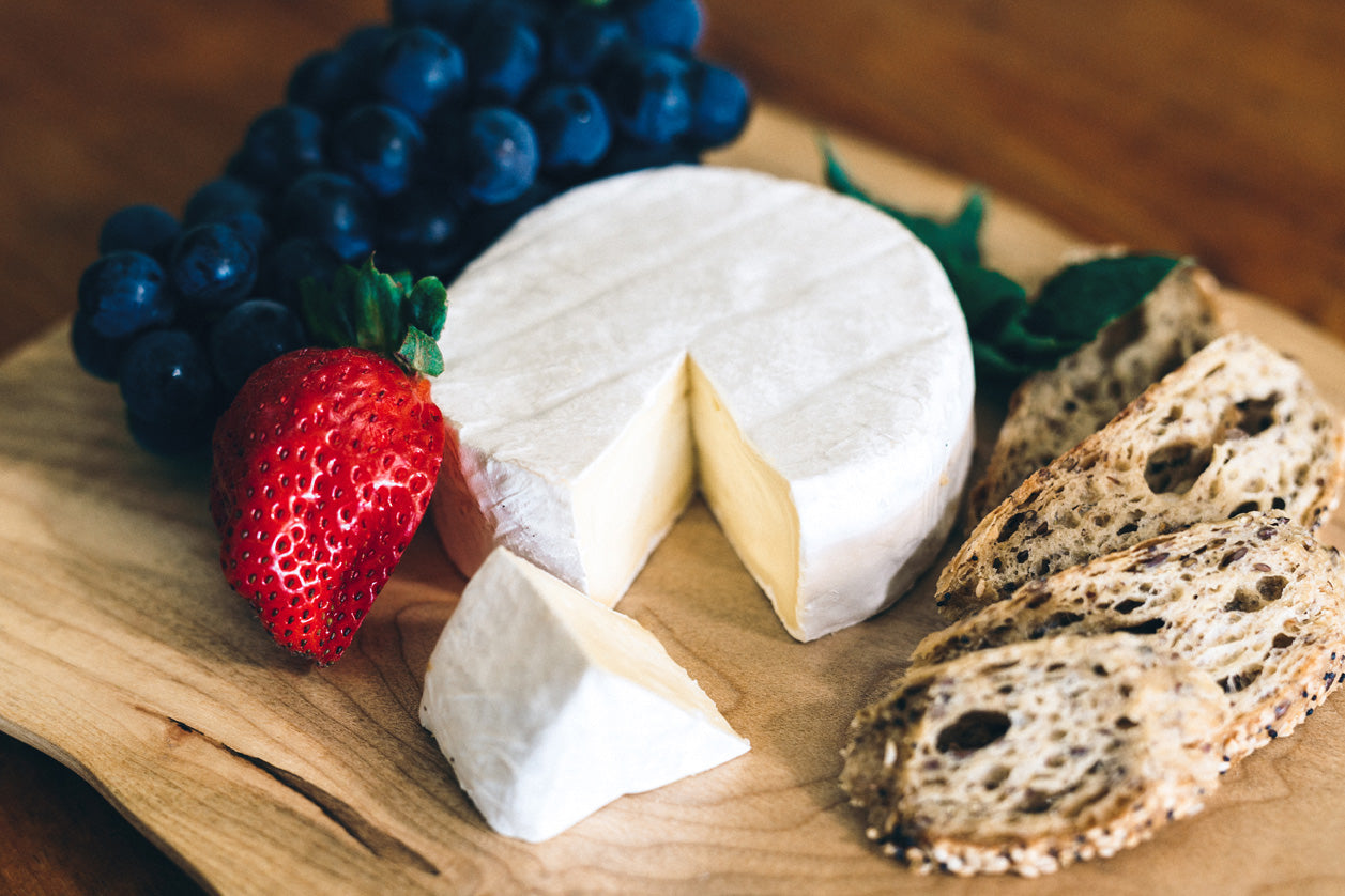Island Brie styled on a wooden board with fresh strawberries, grapes, and slices of rustic bread. A wedge is cut from the wheel of brie, revealing a creamy interior beneath the velvety white rind.