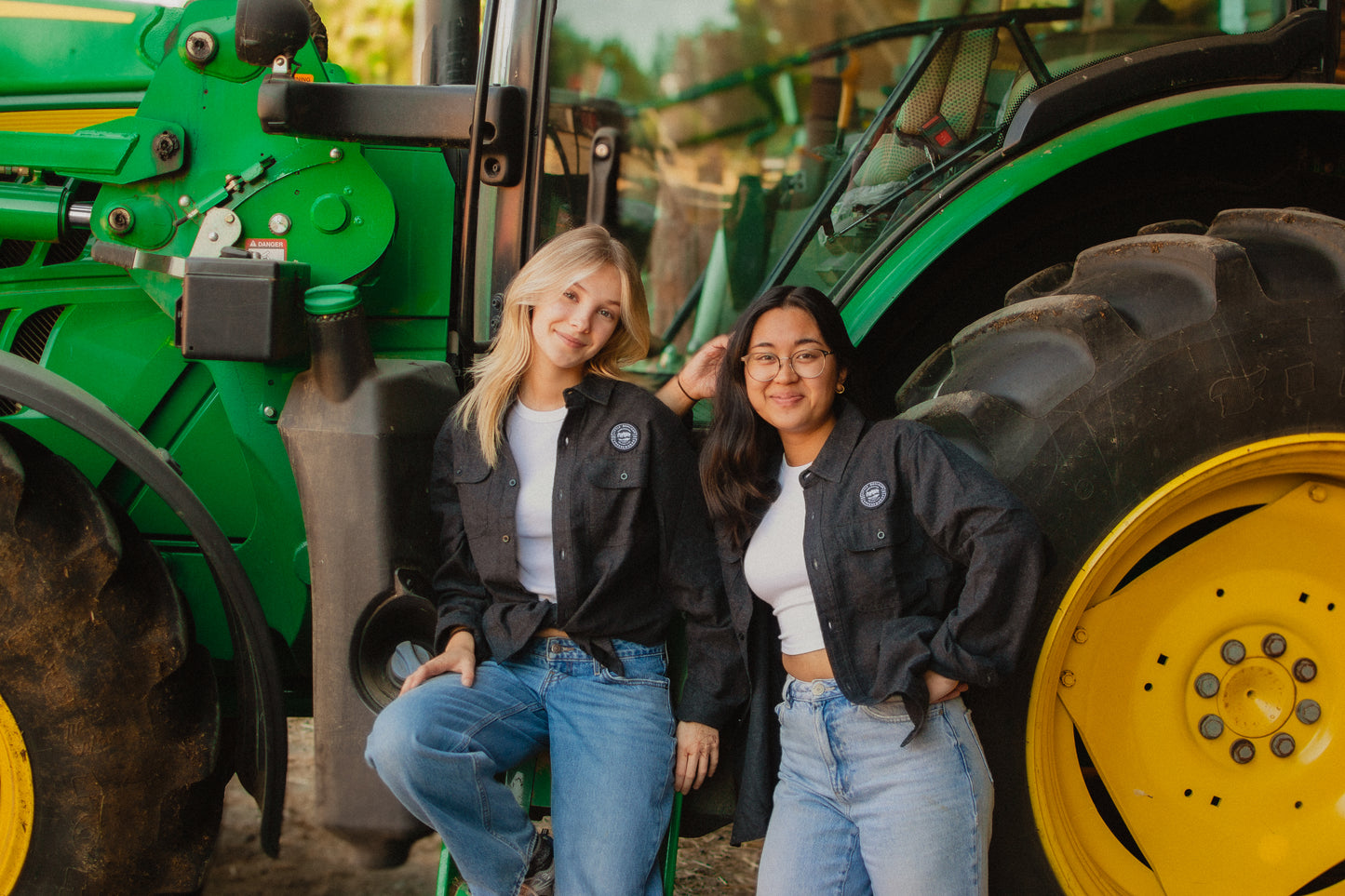 Two people posing in front of a green tractor, wearing Little Qualicum Cheeseworks' flannel patch shirts.