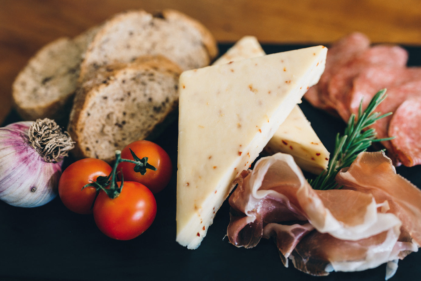Qualicum Spice cheese is displayed with an array of foods, including sliced cured meats, tomatoes, garlic, and sliced bread. The cheese is subtly flecked with red flakes of paprika.