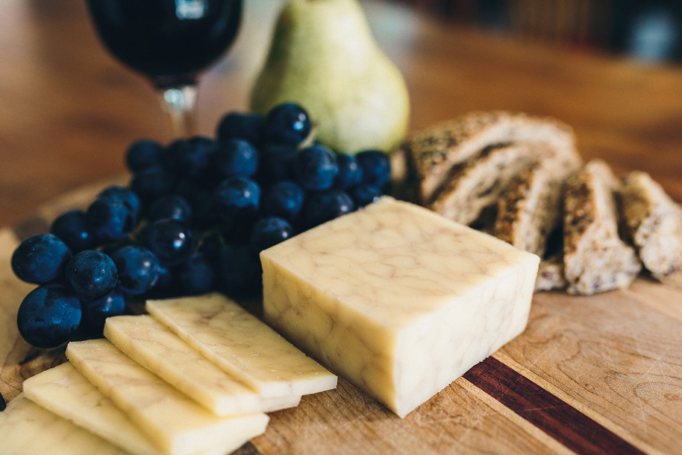 Tipsy Jill cheese is displayed on a wooden board with fresh grapes, sliced bread, and a glass of red wine. The cheese itself has a soft marbling from the addition of red wine.