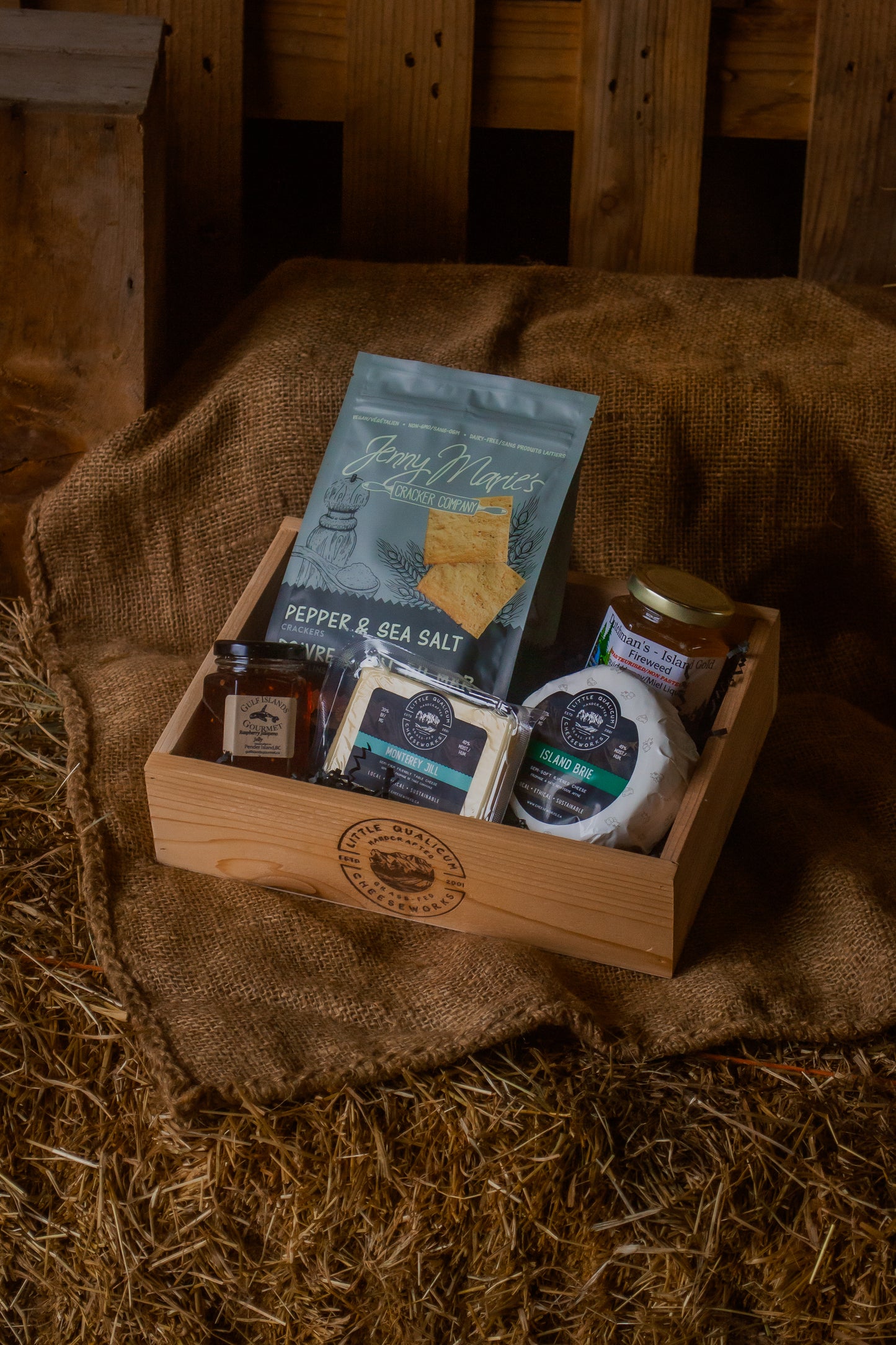 A wooden gift box with various Vancouver Island local items including a jar of jam, honey, crackers, and Brie and Monterey Jill cheese on a rustic wooden background.