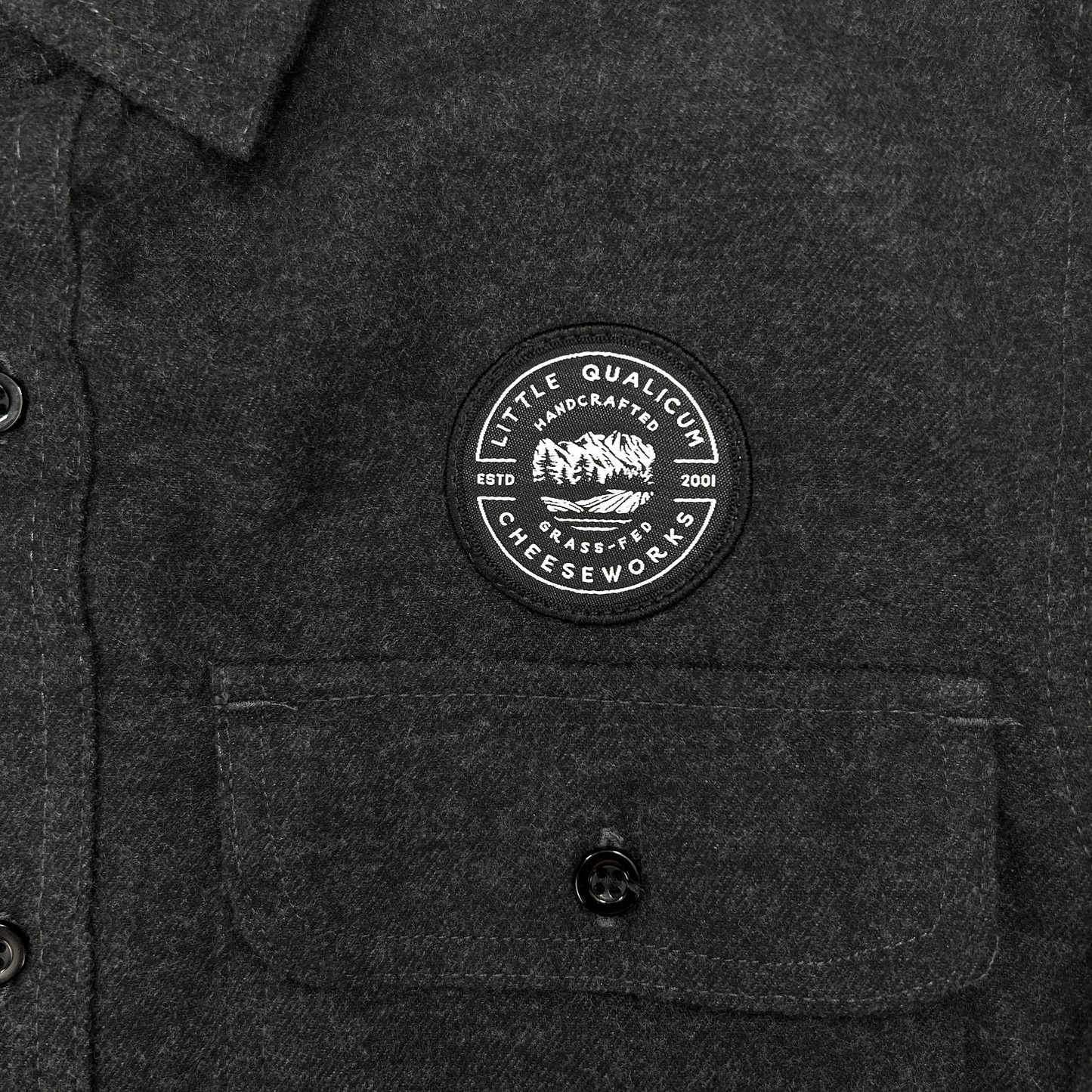 Dark gray shirt with a circular patch on the chest featuring Little Qualicum Cheeseworks' logo (a circular patch withtext and a mountain graphic).