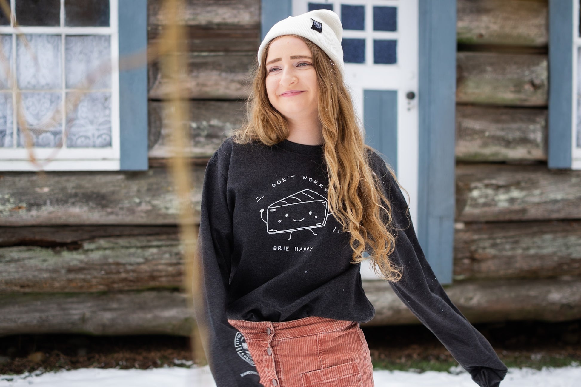 Employee wearing Little Qualicum Cheeseworks toque and don't worry brie happy sweatshirt in front of cabin