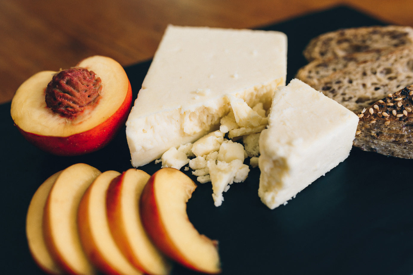 Caerphilly cheese is served on a slate with sliced peach and a seedy bread. The cheese shows a moist crumbly texture.