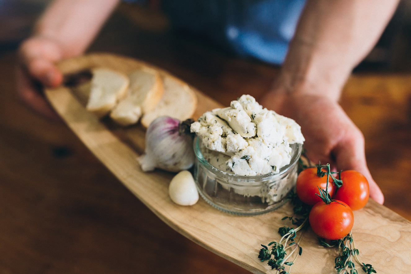Fromage Frais Herbie is styled in a serving jar alongside a head of garlic, herbs, and baguette.