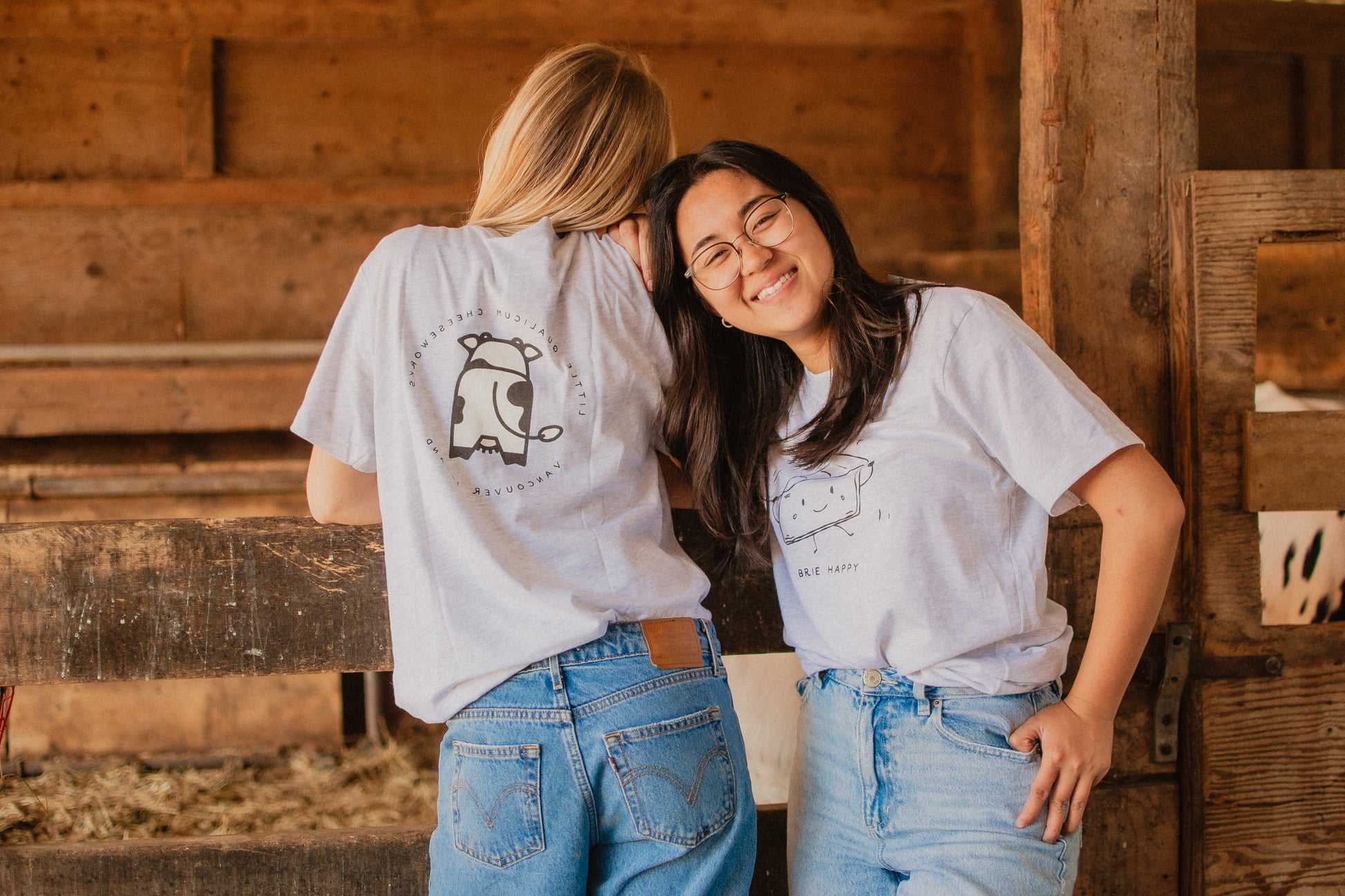 Two people wearing t-shirts with cow and cheese illustrations in a rustic wooden barn.