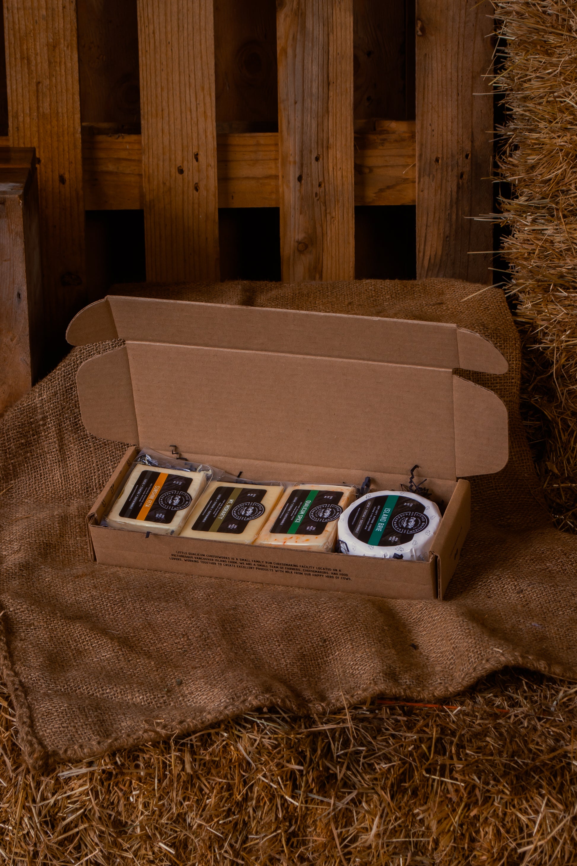 Open cardboard box with brie, qualicum spice, mount moriarty and sunshine feta on a rustic wooden and hay background