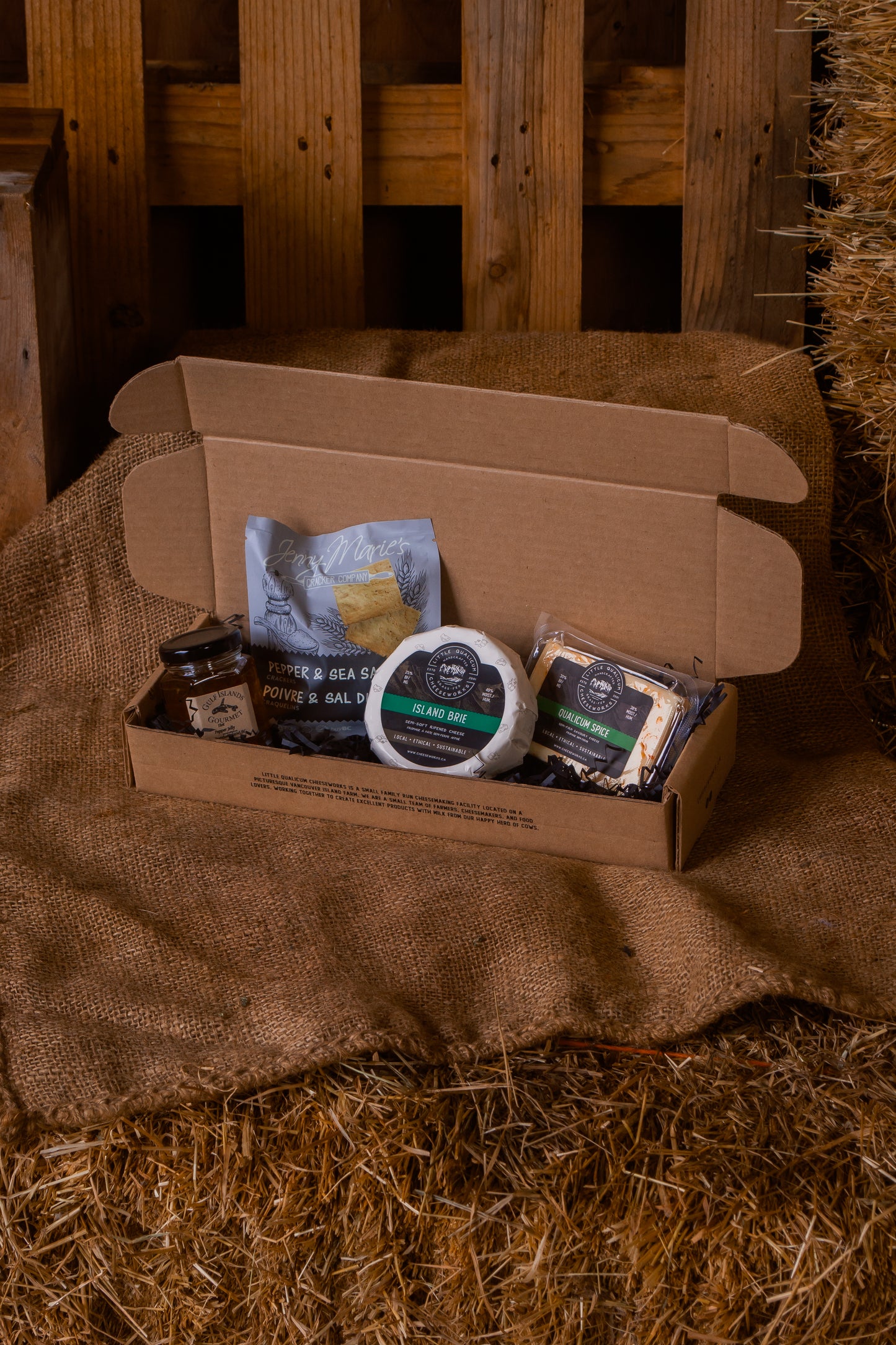 Open cardboard box with brie, qualicum spice cheese, pepper and sea salt crackers and gulf islands jelly on a rustic wooden and hay background