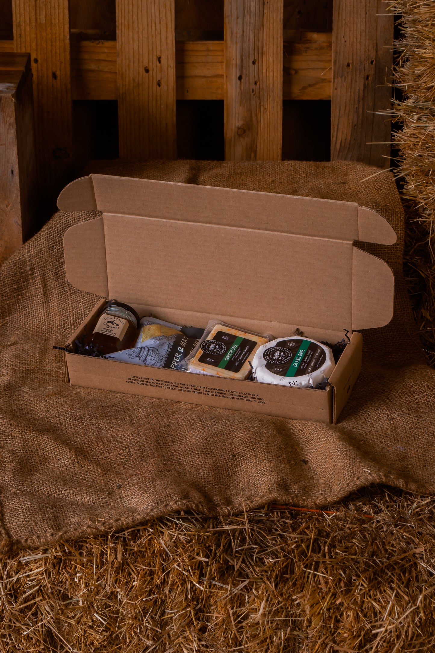Open cardboard box with brie, qualicum spice, pepper and sea salt crackers and gulf islands jelly on a rustic wooden and hay background