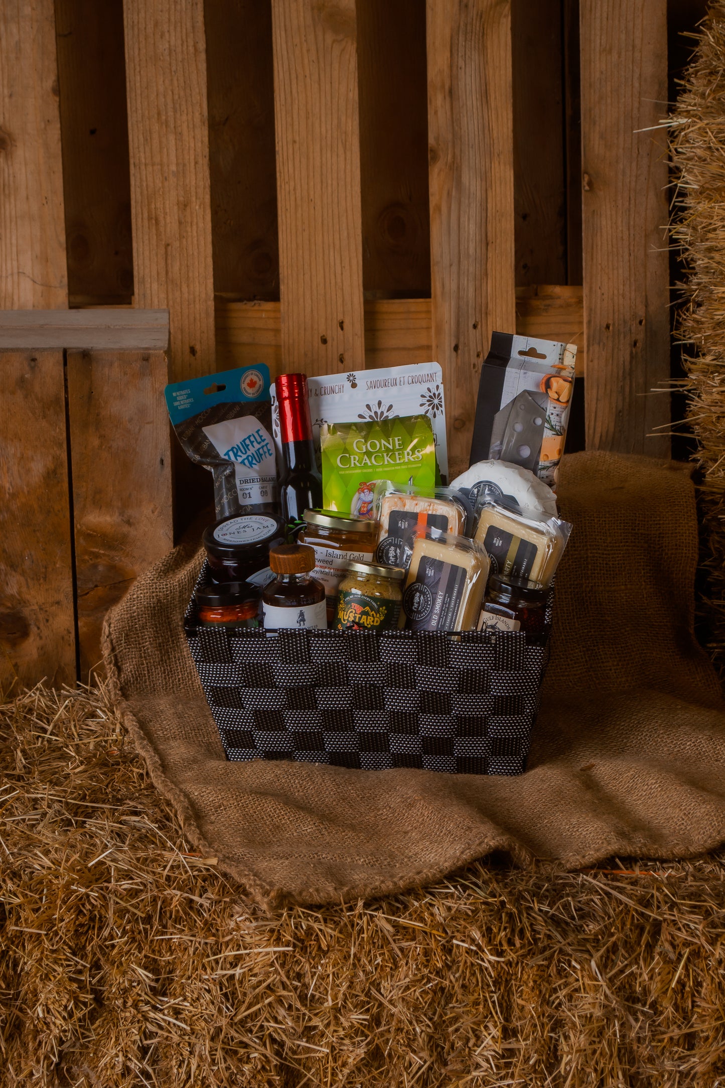 Gourmet gift basket with cheese and various local foods and parings on a hay bale against a wooden background.