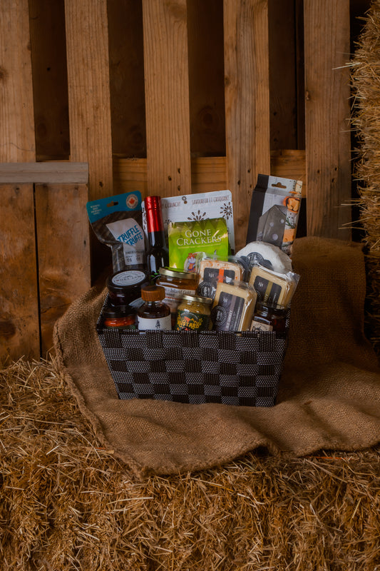 Gourmet gift basket with cheese and various local foods and parings on a hay bale against a wooden background.