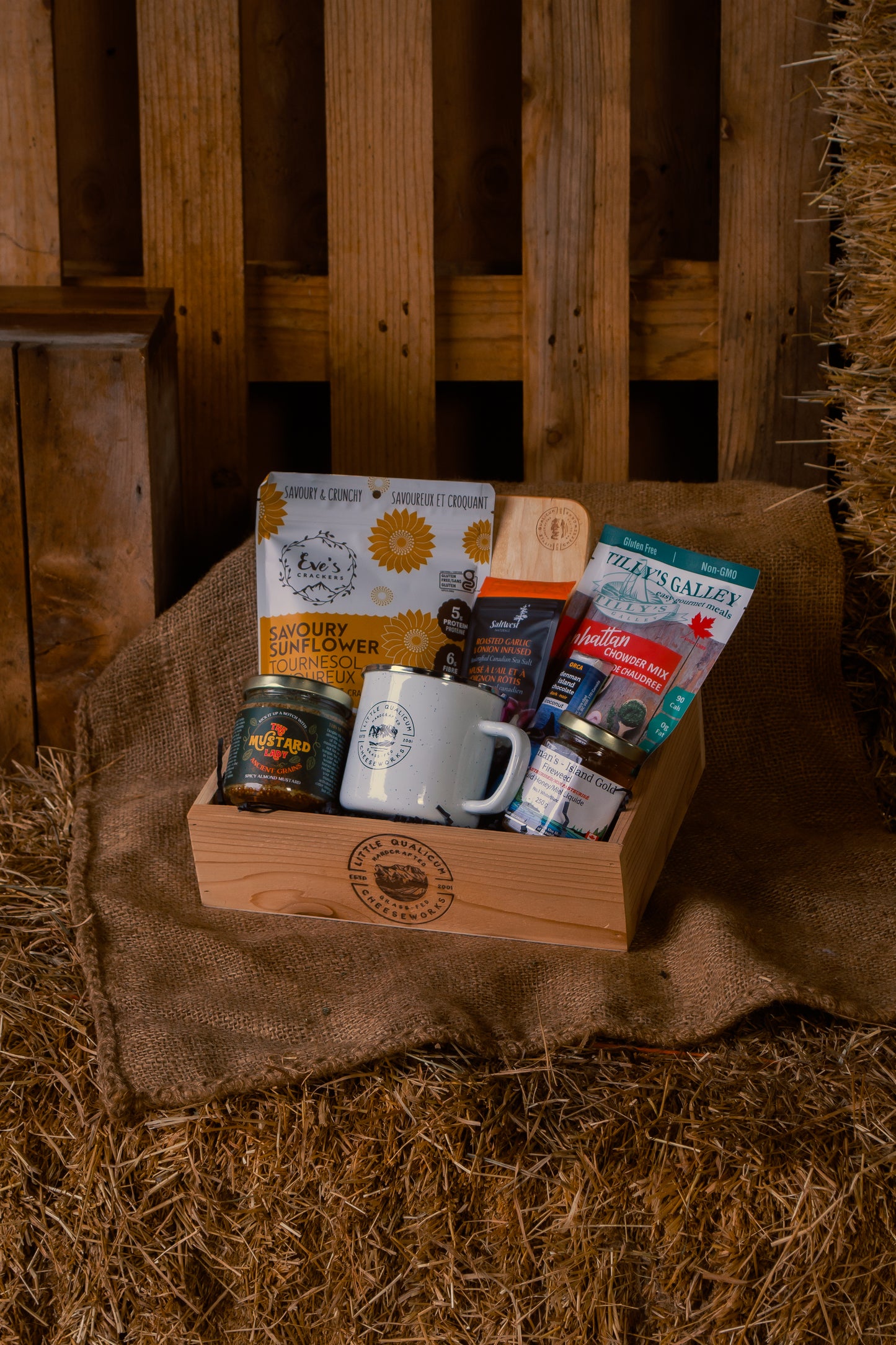 Wooden gift box with various items including a mug, mustard. sunflower crackers. salt, cutting board, soup mix and honey on a rustic wooden and hay background