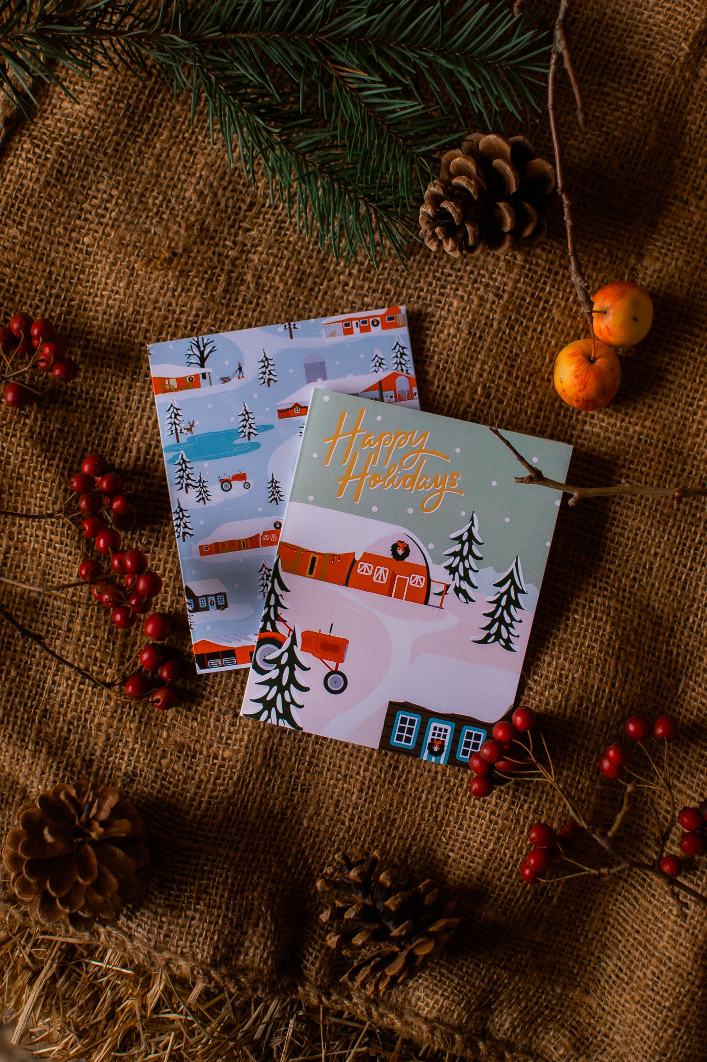 Two Christmas-themed greeting cards on a rustic surface with pine cones and berries.