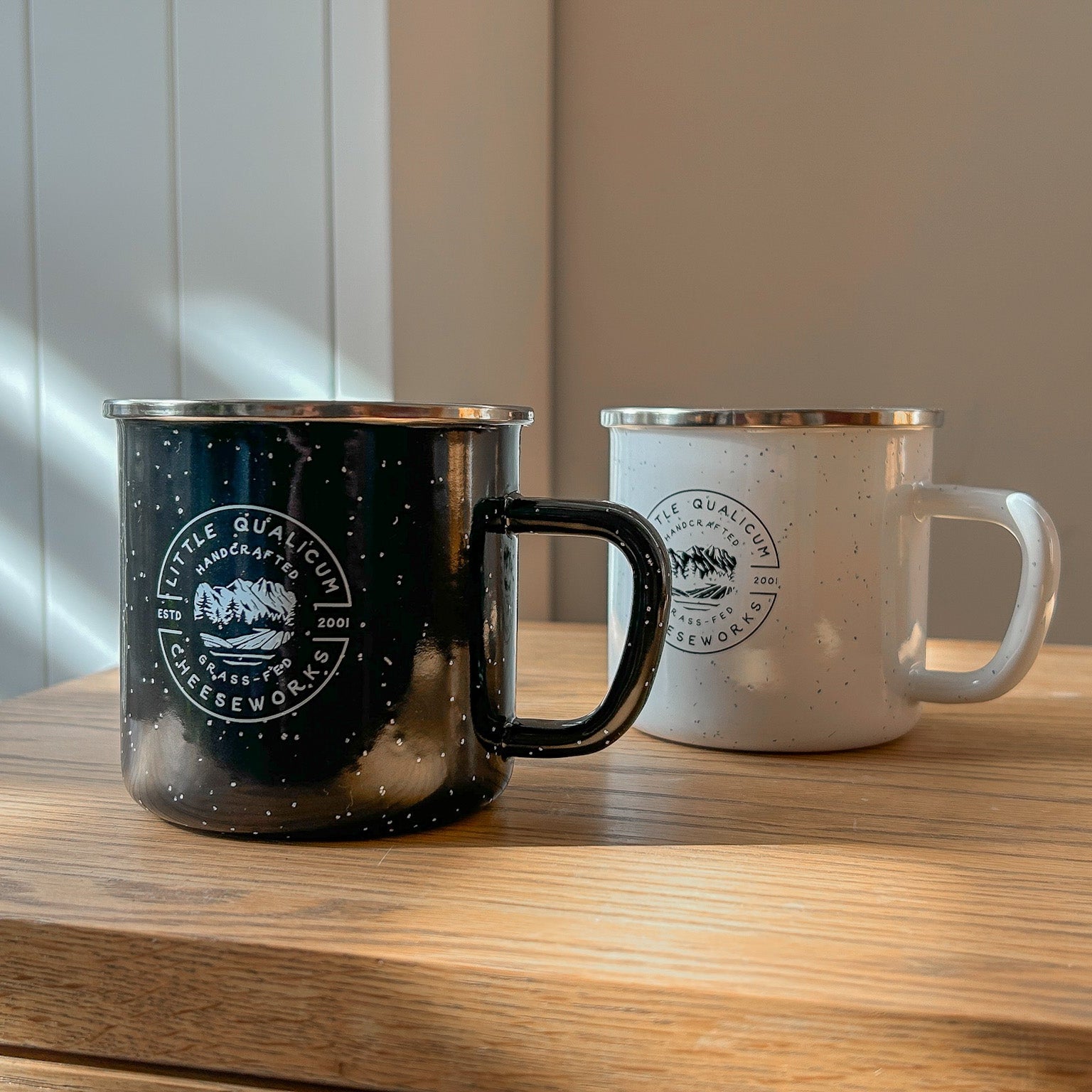 Two camp mugs, one black and one white, with Little Qualicum Cheeseworks' logo, sitting on a wooden table in natural light. The mugs are enameled metal and feature a vintage-style speckled pattern in the finish.