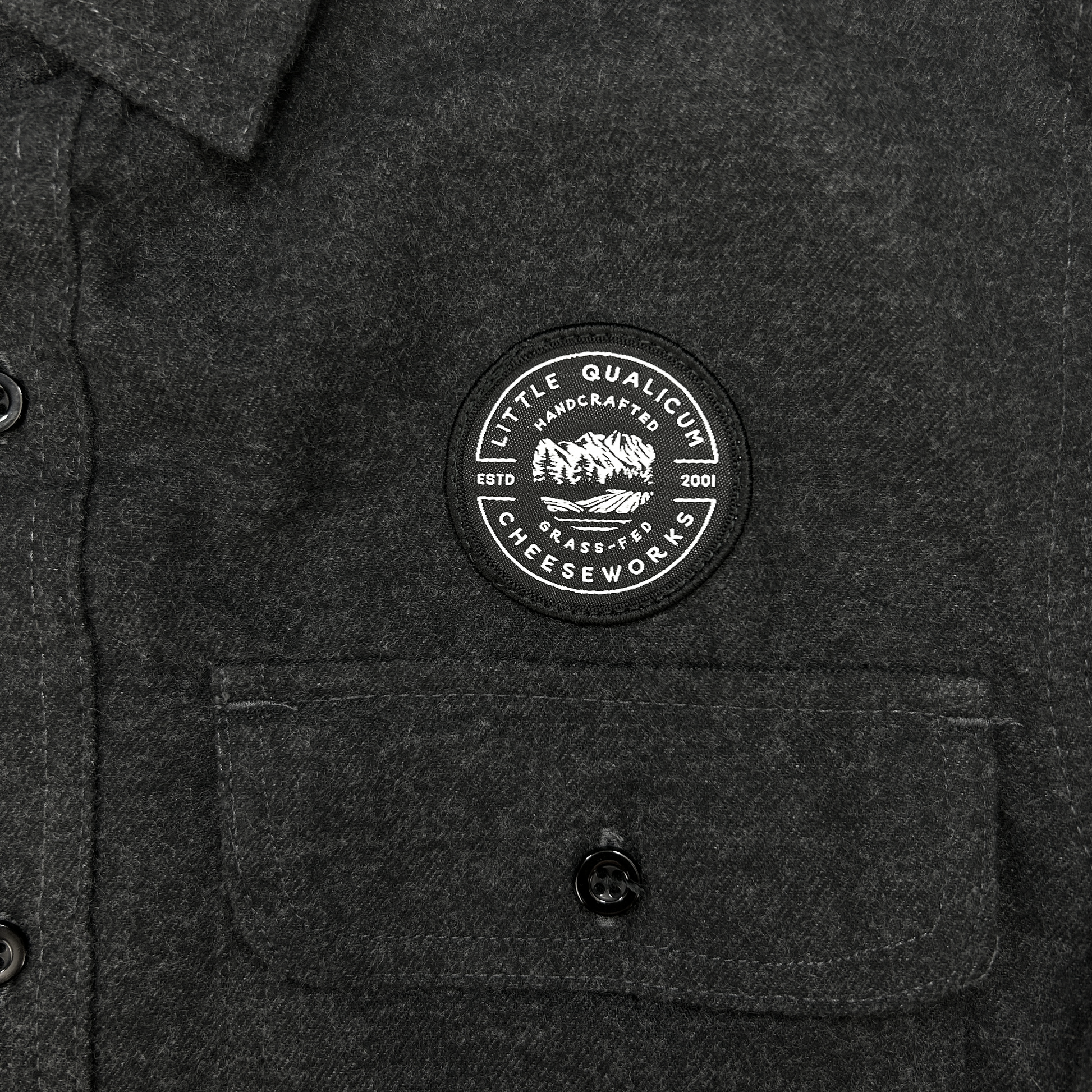 Dark gray shirt with a circular patch on the chest featuring Little Qualicum Cheeseworks' logo (a circular patch withtext and a mountain graphic).